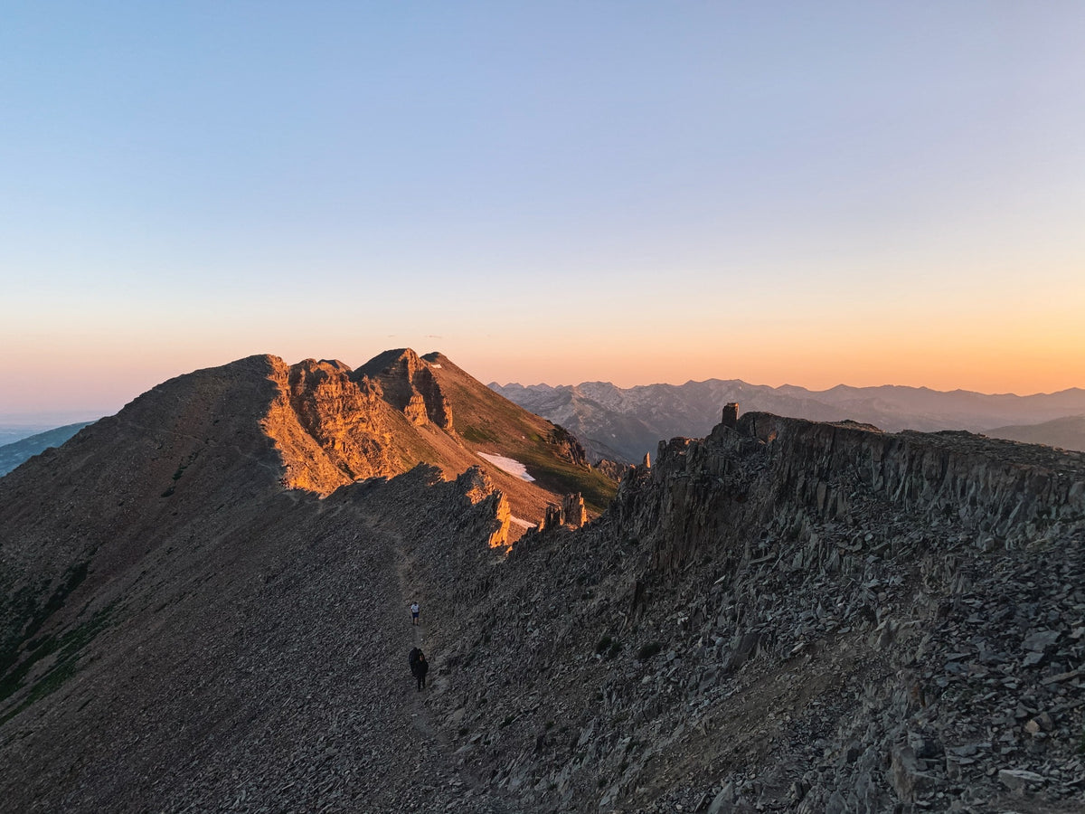 Golden sunlight illuminates a rugged mountain ridge and rocky trail with two distant hikers at sunset